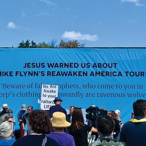 Photo of a cerulean blue billboard with black text that reads "Jesus Warned Us About Mike Flynn's ReAwaken America Tour." Foreground features a group of people facing the billboard; one protestor's sign reads "We Are Awake to Your Traitorous Lies"