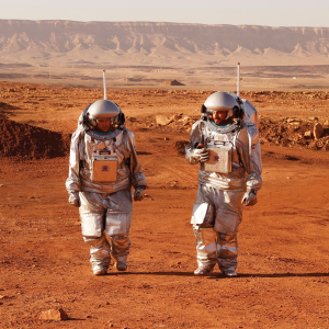 Two people in astronaut suits walk in an orange-brown desert landscape