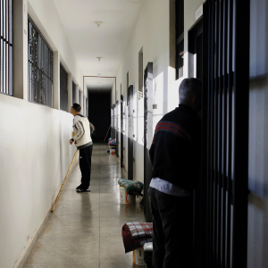 Pictured is a hallway with white walls and barred windows. There is a man in a black pants and a white sweatshirt looking out of the window, with another man standing down the hall looking at him. 