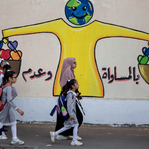 Women wearing hijabs walk with young girls wearing gray school uniforms past a mural depicting a smiling globe and hearts.