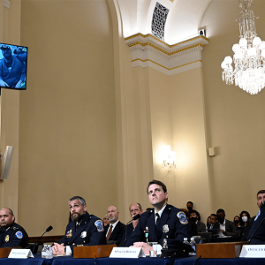 Four men in law enforcement uniforms sit at a table. Behind them, a screen shows protesters on Jan. 6.
