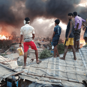 Rohingya youth stand atop tarp roofs as they watch fire spread in the treeline