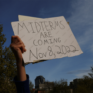 A demonstrator holds a sign reading "Midterms Are Coming Nov 8, 2022" against a blue sky.