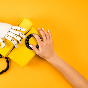 A human hand and a robotic hand both reach for a vintage telephone on a yellow background.