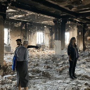 Three people walk amid rubble in a damaged building with a ceiling that is blackened.