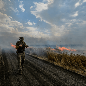 A Ukrainian serviceman holds a gun while walking through a burning wheat field.