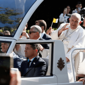 Pope Francis wears white robe and stands in an open car surrounded by a crowd.