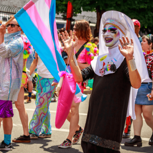 A person wearing heavy drag makeup and a black Catholic habit waves in a pride parade.