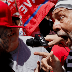 A closeup of two men yelling at each other. One is wearing a red hat, the other is in a black and white striped prison suit. 
