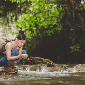 A woman wearing jeans and hiking boots kneels in the forest splashing water on her face