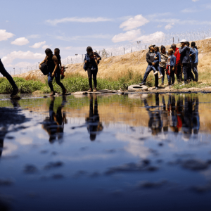 Shadows fall over migrants who are crossing a river. Their reflections appear in the surface.
