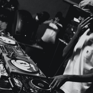 A man wearing a white t-shirt has one hand on his headphones and the other on a turntable.