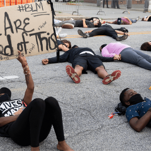 Protesters lie on pavement. One holds a sign that says "We Are Tired of This."