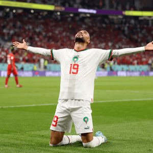 A soccer player kneels on the field with arms outstretched. Behind him, fans are crowded in stadium seating.