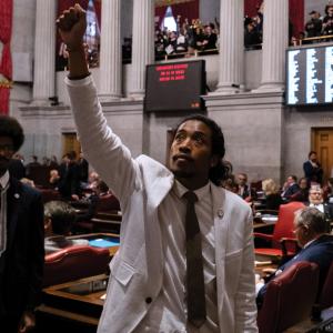 Representative Justin Jones, a Black-Filipino man in a white suit and brown tie, stands amid the aisles of Tennessee's House of Representatives, raising his fist to the ceiling as his colleague Justin Pearson stands behind him to the left.