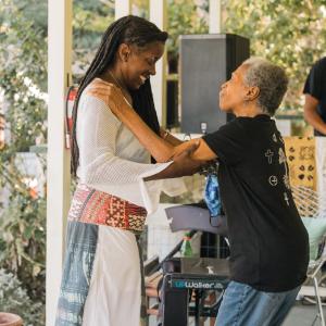 A black woman in a white dress with long dark brown hair smiles while dancing with her elderly mother on a porch, who's wearing a black t-shirt and jeans. A man in a black t-shirt plays music in the background with a laptop and speaker on a table.