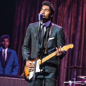 Garrett Turner is dressed in a black suit playing an electric guitar as Ike Turner in the musical 'TINA: The Tina Turner Musical.' A black man in a blue suit is playing the keyboard in the background, where both men are flanked by a purple stage curtain.