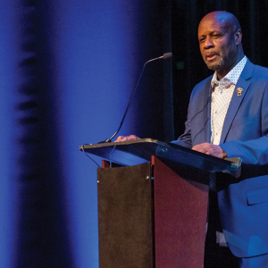  A Black man in a royal blue suit with a white collared shirt stands at a podium; background of the image is dark, with two vertical stripes of blue light extending top-to-bottom to the left of the man.