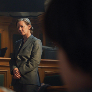 The photo shows a woman with gray hair in a gray suit in a courtroom, looking at people off camera. 