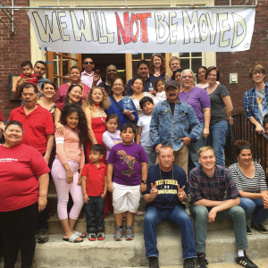 The photo shows a large group of people smiling for a photo underneath a banner that reads "We Will Not Be Moved." The people are standing in front of the door to their apartment complex. 