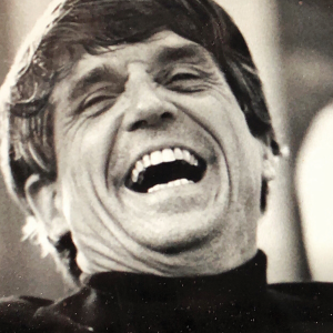 A white man wearing a black turtleneck crinkles his face with exuberant laughter