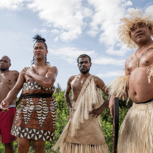 The photo shows four men from the Pacific Islands, some are in traditional dress made of straw and other natural fibers, and one of the men is holding a flag that is wrapped around the flag pole