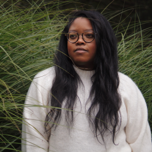 Cole Arthur Riley, a Black woman wearing glasses and a white sweater, stands before tall grasses