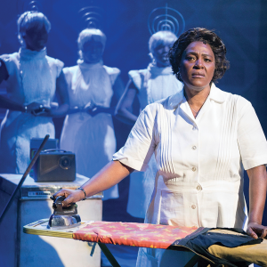 A weary middle-aged Black woman looks up from ironing clothes in a dark room