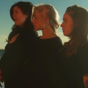 From left to right, musicians Lucy Dacus, Phoebe Bridgers, and Julien Baker are dressed in black and cast in the warm glow. They stand in a cascading line next to one another, staring off beyond the left side of the photo with waves in the background.