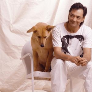 A photo of Rich Mullins in a white t-shirt and white pants. He's sitting on a white chair with a white sheet draped in the backdrop. He's leaning forward on his knees with a grin on his face. A dog with tan-colored fur sits to his right on the same chair.