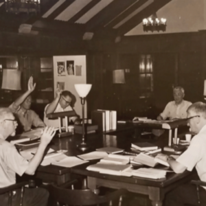 The image is a black and white photo showing a group of old white men sitting around a table with Bibles and other documents, some have their hands raised. 