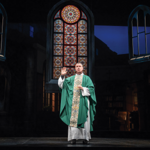 The image is a picture of a priest on a stage with a green robe, with a stained glass window in the back