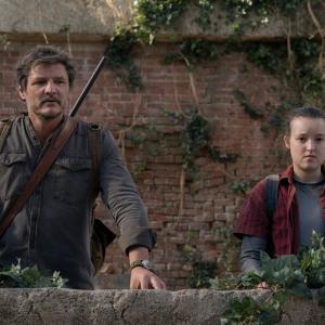 Joel (actor Pedro Pascal) and Ellie (actress Bella Ramsey) from 'The Last of Us' HBO series are standing side by side on the roof of a neglected building with their arms leaning on a brick wall covered in foliage.