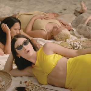 Lorde, wearing yellow, sings to the camera as she lays on a blanket on the sand