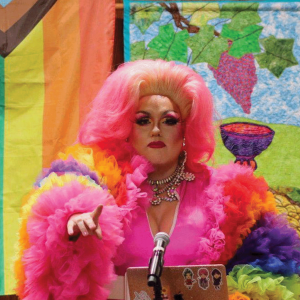 A person wearing a tall pink wig and a pink dress with rainbow fluffy sleeves is standing at the pulpit of a church, preaching. There is a pride flag in the background. 