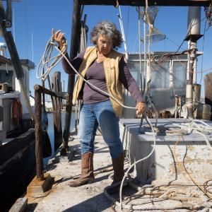 The author of this piece, Diane Wilson, is wearing a tan vest with a purple long-sleeve shirt, jeans, and brown boots. She's rolling up rope on a boat, which is docked next to another one to the left.