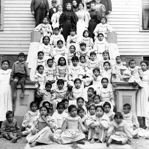 An old black-and-white photo of students and teachers sitting and standing on the steps of the Thomas Indian School building in the 1890s. 