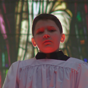 A young boy wearing a white surplice stands before a wall of stained glass