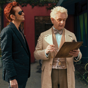 The photo shows two men, one who is an angel and dressed in lighter colors, and another who is a demon dressed in black. The angel is looking at a clipboard and the demon is just standing there. 