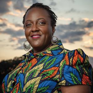 A photo of Heather McTeer Toney: a black woman with short hair, golden circular earrings, and a shirt with a pattern of leaves in vibrant blues, oranges, and yellows. She is looking at the viewer and smiling with a forest and evening sky behind her.