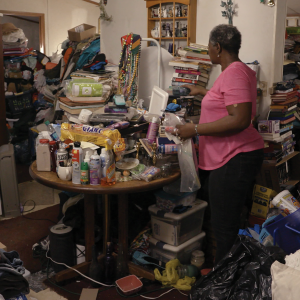 A woman stands in a kitchen with objects piled all over a table and along the walls 