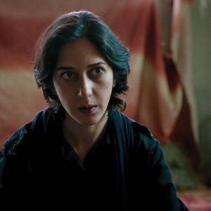 A photo of actress Zar Amir Ebrahimi as fictional journalist Arezoo Ramimi in the film 'Holy Spider.' She is cast against a red flag in the background and staring just off camera at something.