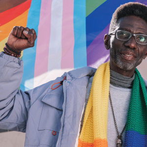 The image shows an older Black man wearing a knitted rainbow scarf and holding up a fist in front of a colorful pride flag. 