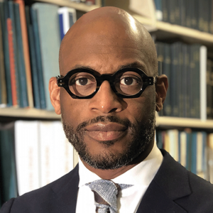 The picture shows a bald Black man with facial hair wearing black glasses in front of a bookshelf, wearing a suit. 