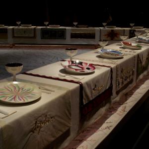 A picture of Judy Chicago's art exhibit called "The Dinner Party." There are unique plates, glasses, silverware, and tapestries depicted for esteemed women across time (mythological and historical) around a triangular banquet table. 