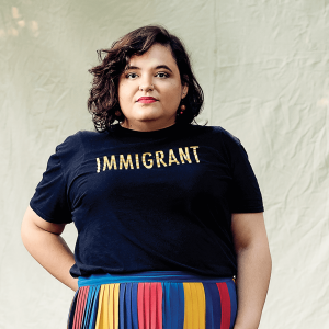 Aline Mello stares seriously at the camera wearing a multicolored skirt and a t-shirt that says "Immigrant"