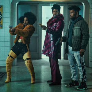 The picture shows three black youth, dressed in colorful outfits and pointing handguns at something off screen. 
