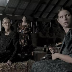 A group of Mennonite women are standing and sitting in a barn filled with crates and hay bales in the film 'Women Talking.'