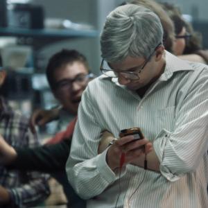 A photo from the docufilm ‘Blackberry.’ Actor Jay Baruchel is Mike Lazaridis, co-CEO of Blackberry. He has short gray hair, glasses, and wears a white dress shirt. He glares down at a phone with wires plugged into it. People behind him are cheering.