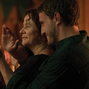 A son embraces his mother from behind, who lift up their hands together to clap.